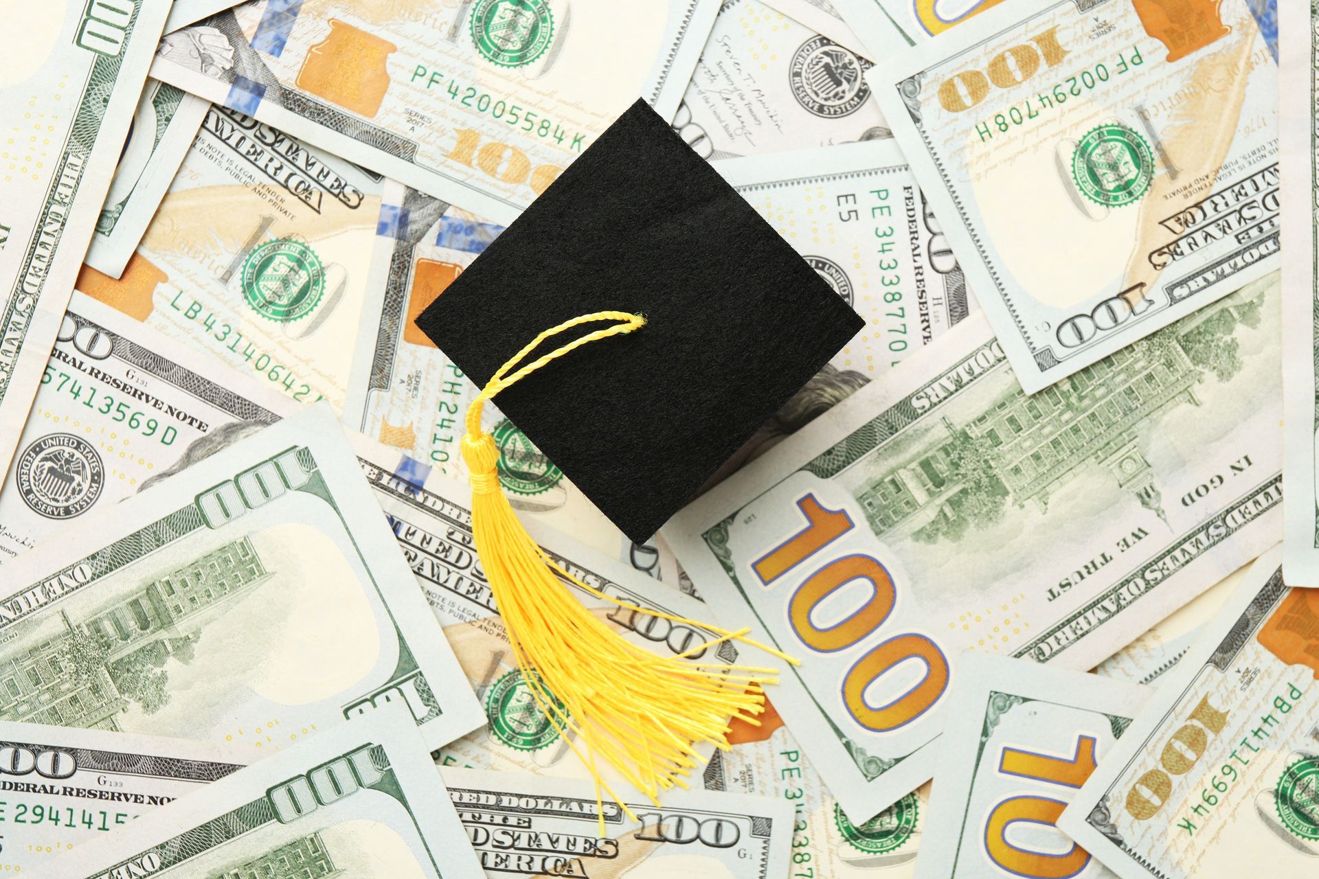 Black graduation cap with yellow tassel on a scattered bed of hundred-dollar bills.