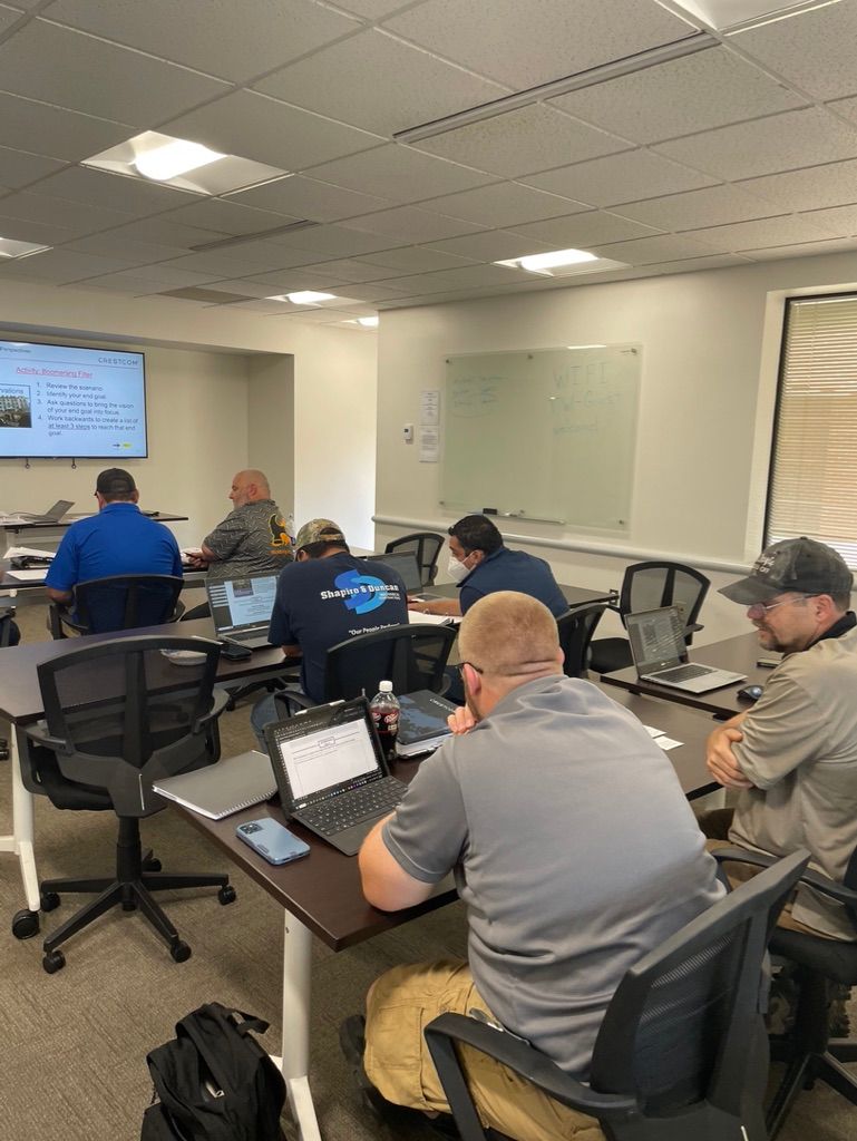 A group of people in a classroom, attending a training session with laptops and a projector screen.