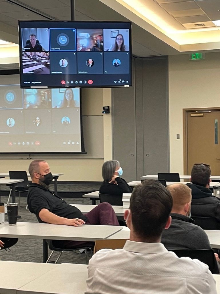 People in a classroom watch a video conference on a screen. Some wear masks, and one man reclines on a desk.