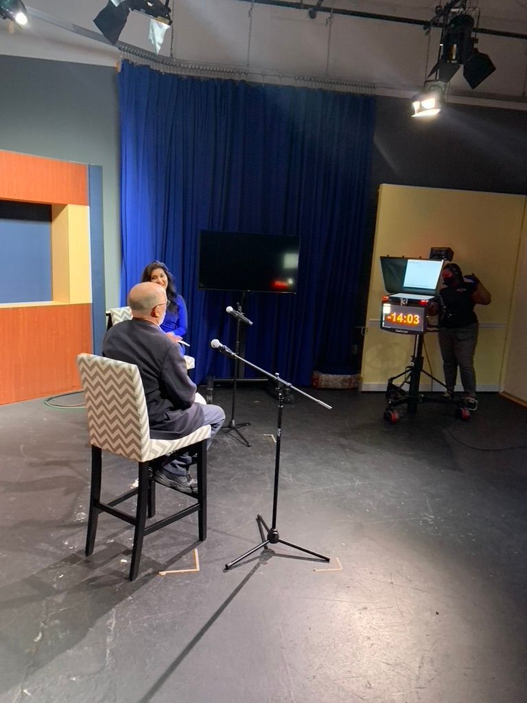 Two people sit in a studio with microphones. A TV screen and camera are also present.