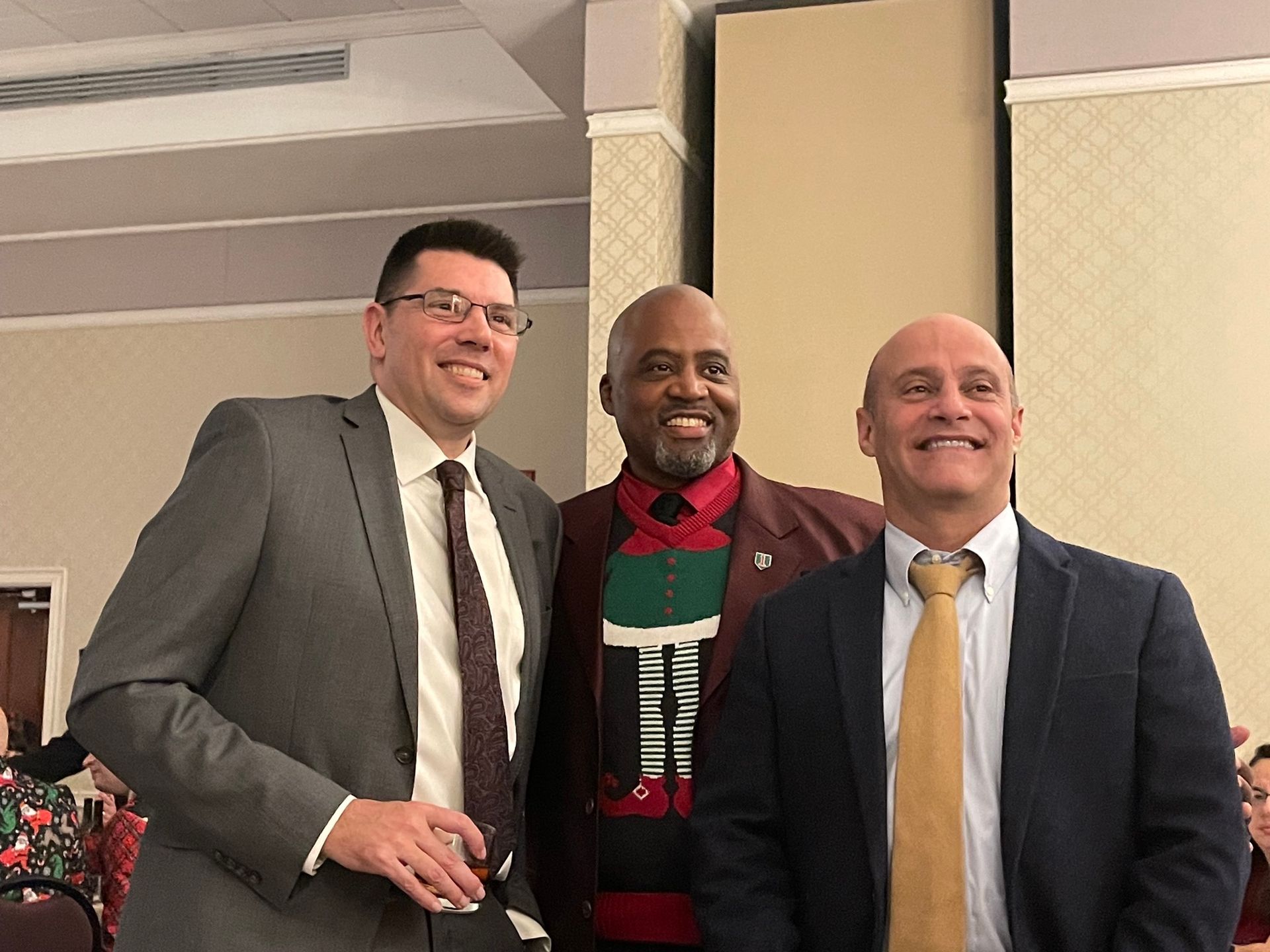 Three men smiling at a social event. Two in suits, one in a festive sweater. Neutral background.