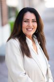 Woman in a white blazer smiling outdoors against a softly blurred background.