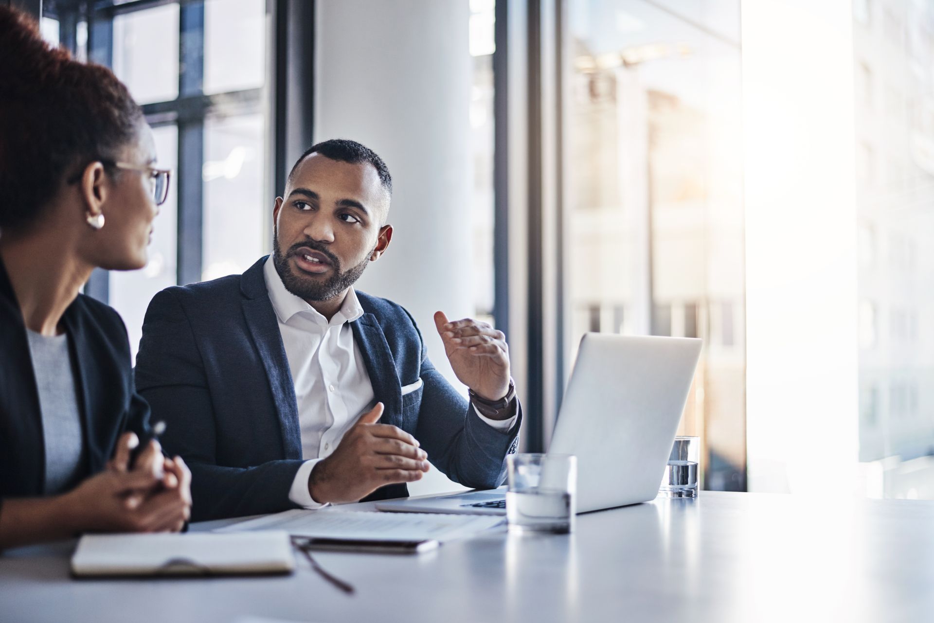 Two coworkers discussing at a conference table with a laptop in a bright office