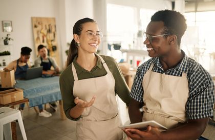 Two people in aprons talking and smiling in a bright workshop, one holding a notebook