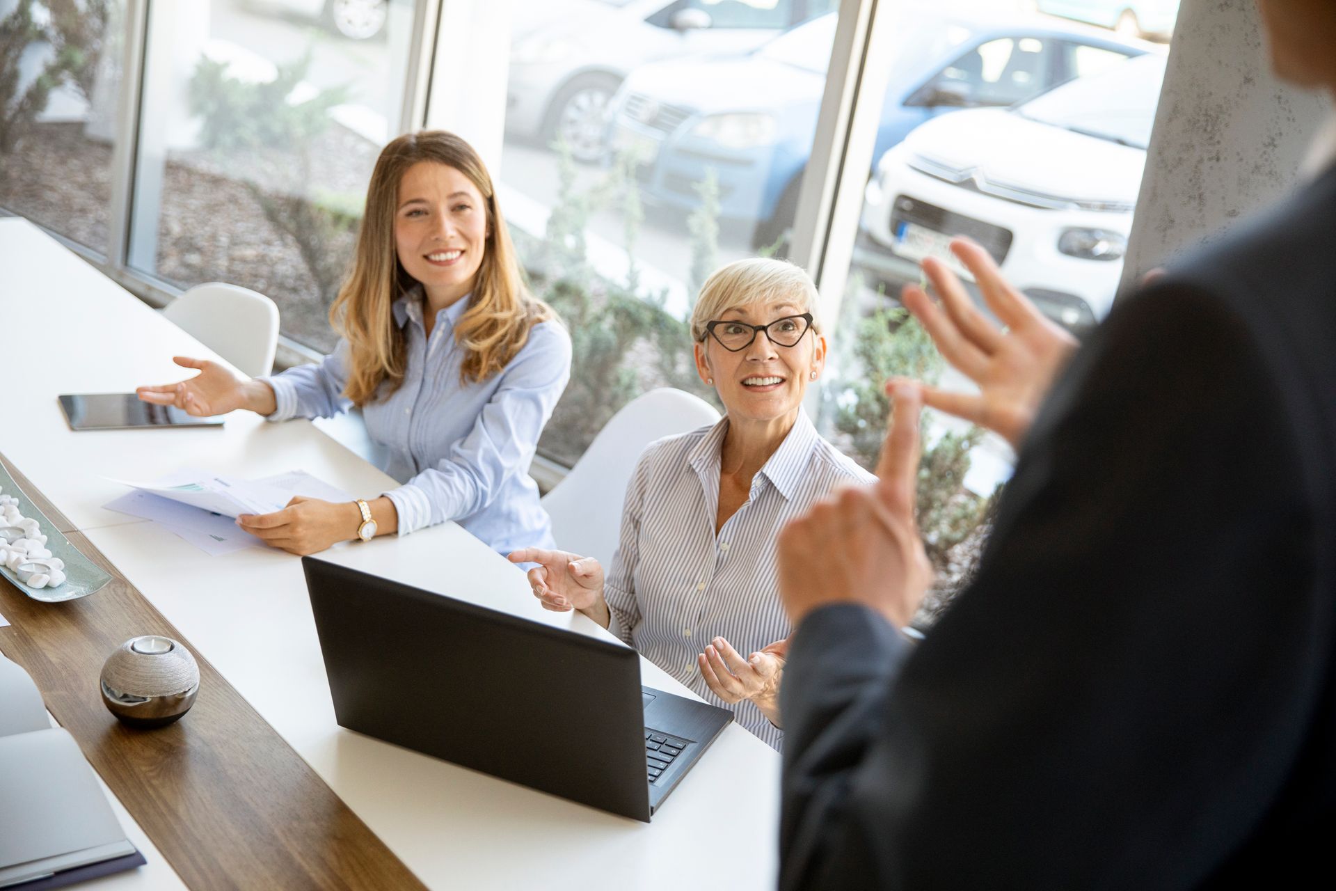 Business meeting in a bright conference room, with two colleagues smiling at a table while another person gestures.