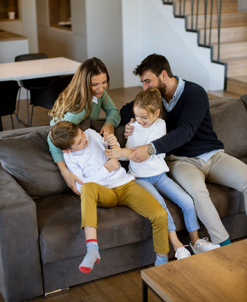 Family of four sitting on a couch, smiling and playing together in a living room.