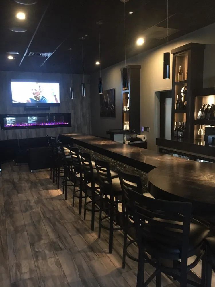 Dim lounge bar with long black counter, high stools, wall TV, and dark wood flooring.