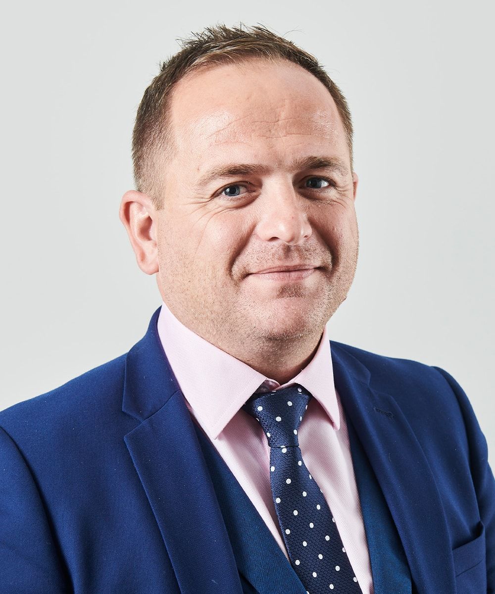 Man in blue suit, pink shirt, and patterned tie, smiling against a gray background.