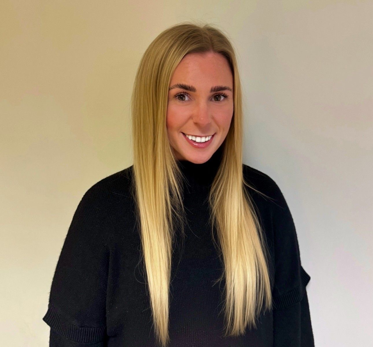 Woman with long blonde hair, wearing a black turtleneck, smiling against a neutral wall.