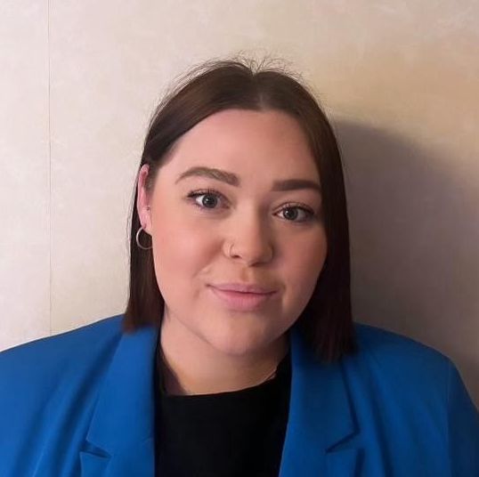Woman with short brown hair, wearing a blue blazer and black shirt, looking at the camera.