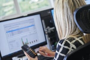 A woman is sitting in front of a computer holding a calculator.