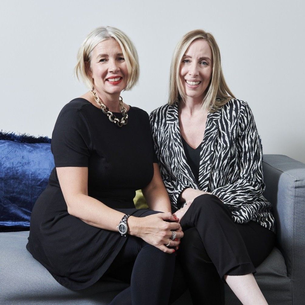 Two women seated on a gray couch, smiling at the camera. One wears a black dress and gold necklace, the other a black and white blazer.