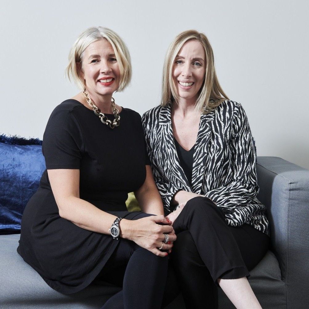 Two women smiling on a gray couch; one in black dress, one in a zebra-print jacket.
