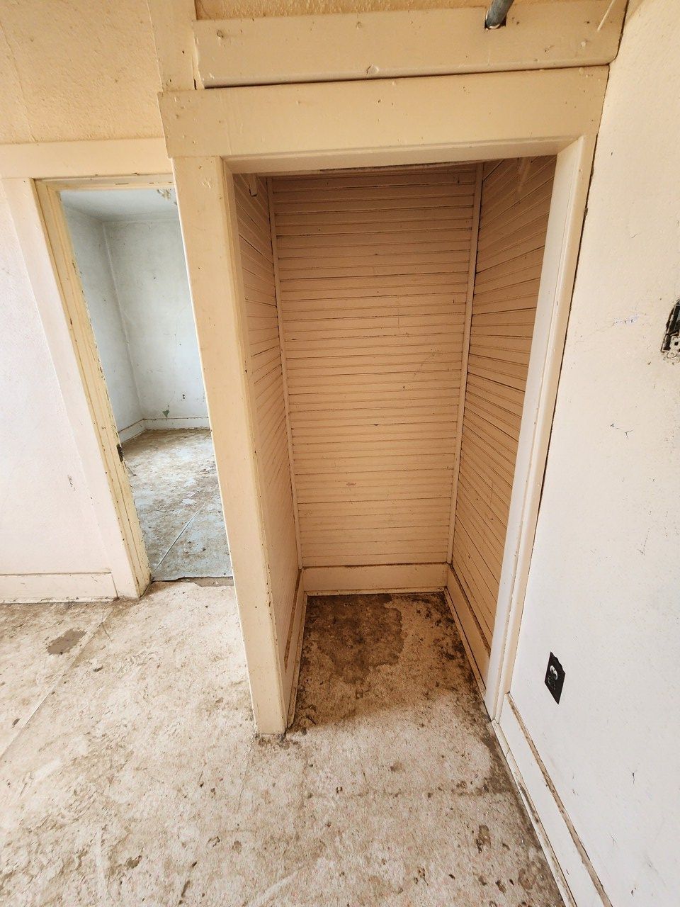 Empty built-in closet with beige slatted back wall and dusty floor in a worn interior room.