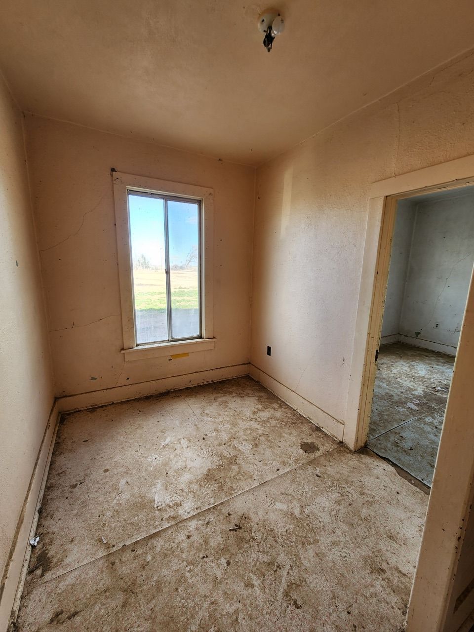 Small empty beige room with dirty floor, window, and open doorway to another room. Small empty beige room with dirty floor, window, and open doorway to another room.