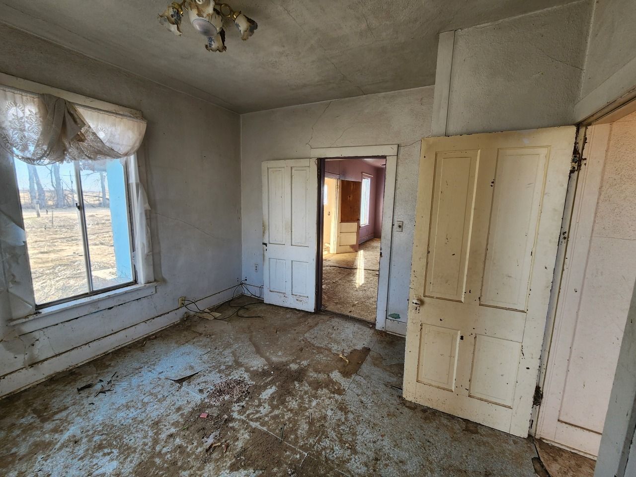 Abandoned room with peeling white walls, dirty floor, and open doorways leading to a sunlit hall