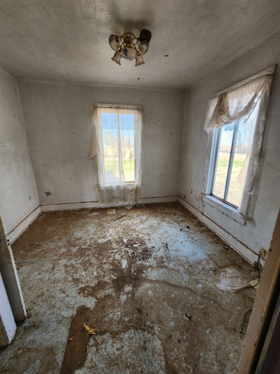 Small empty, deteriorated room with peeling floor, white walls, and two windows letting in daylight