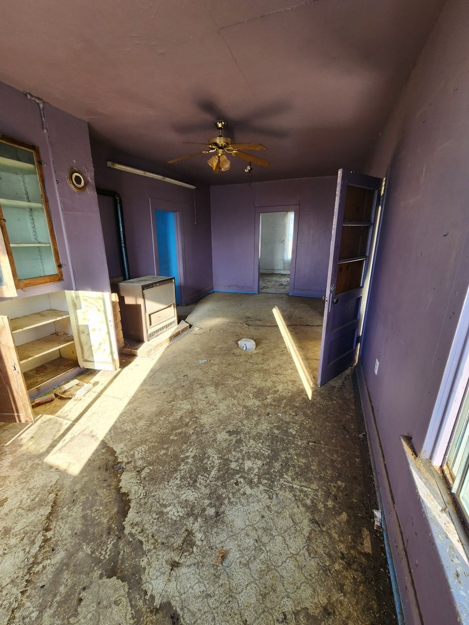 Empty purple living room with worn carpet, fireplace, ceiling fan, and daylight from a window and doorway.