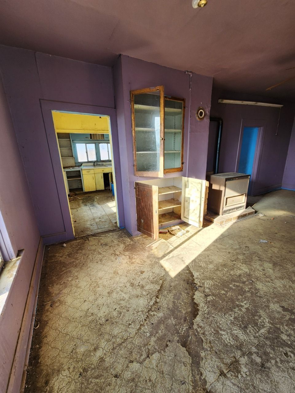 Empty purple room with worn floor, glass cabinet, and sunlight streaming through the doorway