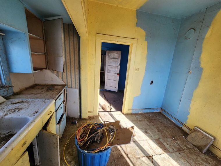 Abandoned kitchen with yellow and blue walls, worn cabinets, debris on the floor, and a doorway to another room