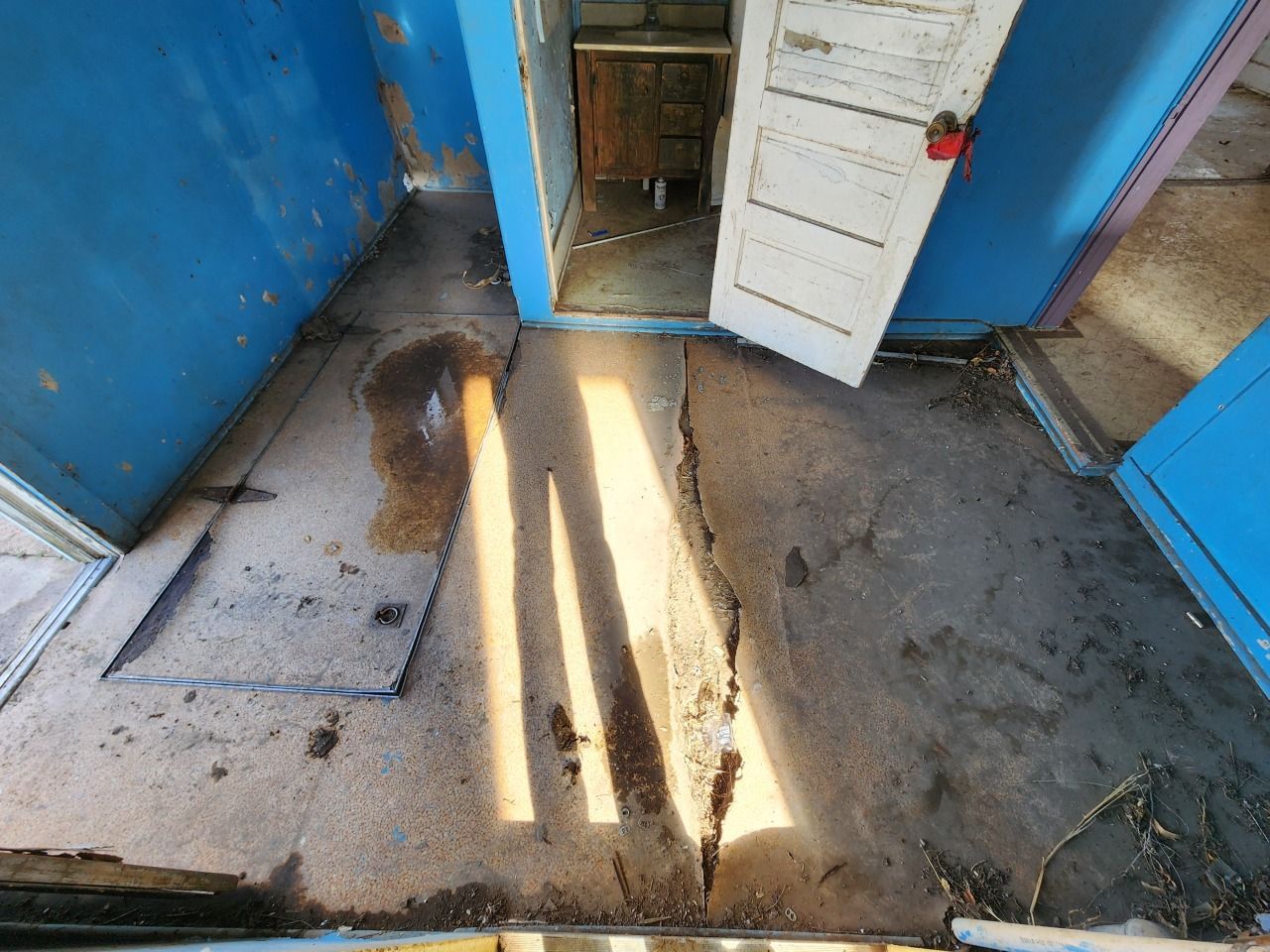Dim blue hallway with open white door, sunlight on worn concrete floor, and a utility hatch on the left. Dim blue hallway with open white door, sunlight on worn concrete floor, and a utility hatch on the left.