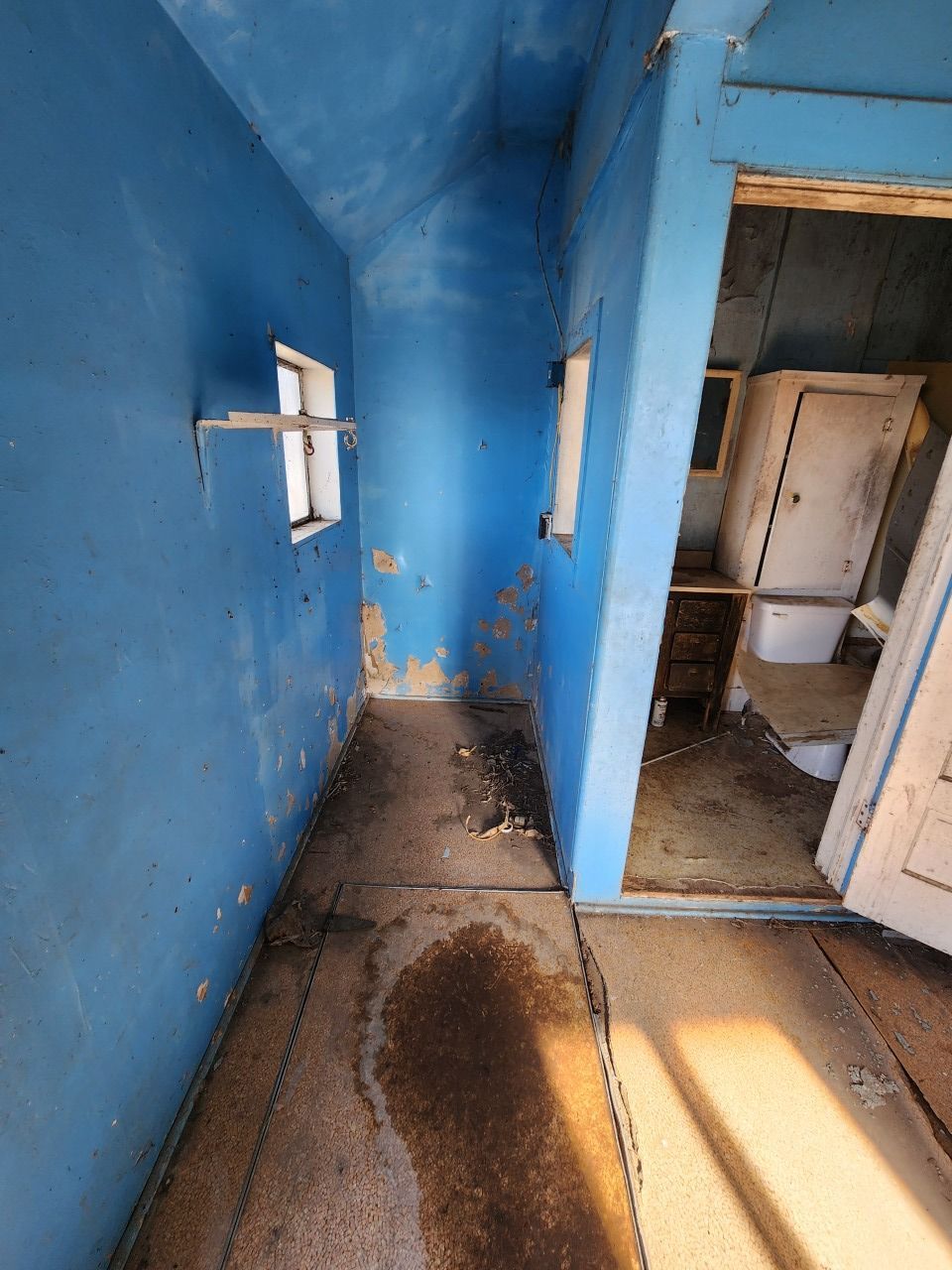 Blue-painted, worn room with damaged walls, a dark stain on the floor, and a doorway to a small bathroom.