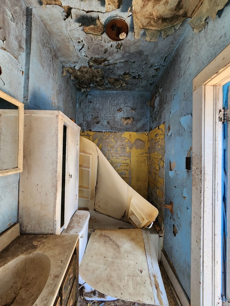 Decayed bathroom with peeling blue walls, broken ceiling, and debris piled around a sink and toilet area