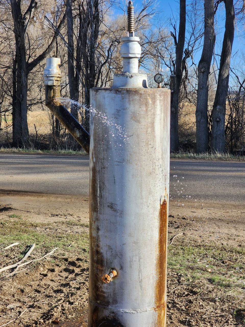 Metal cylindrical tank with a valve, standing outdoors in a grassy yard with leafless trees.