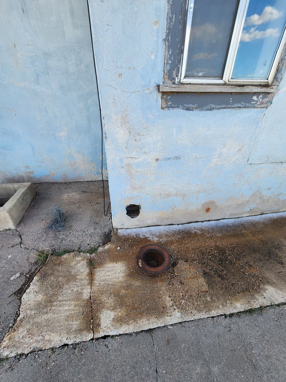Blue exterior wall with a small window and a rusty drain pipe over a stained concrete sidewalk. Blue exterior wall with a small window and a rusty drain pipe over a stained concrete sidewalk.