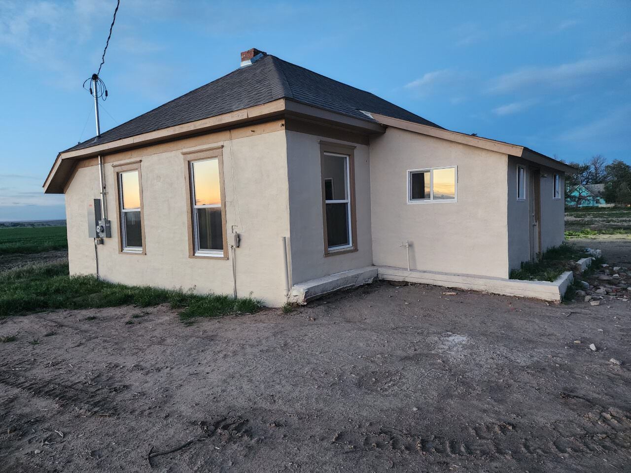 Small beige house with a dark roof at dusk, seen on a dirt lot under a blue sky