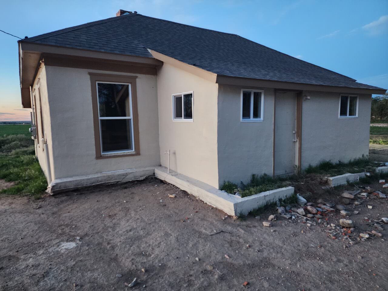 Small beige house with a dark roof and white-trimmed windows on a dirt lot