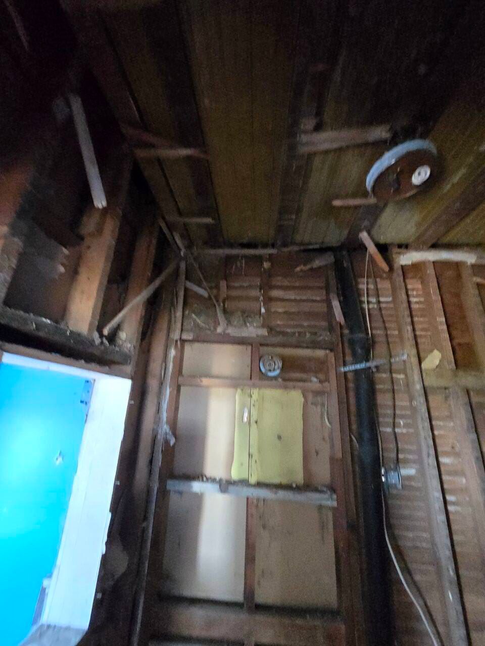 Damaged interior wall with exposed wooden studs, brick, and a small door in a dim room