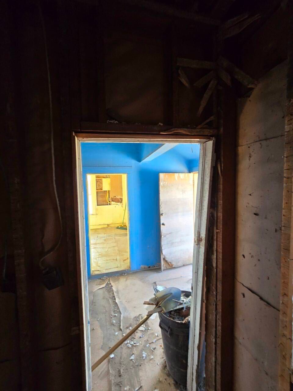 Dark unfinished doorway looking into a bright blue room with a concrete floor and scattered debris