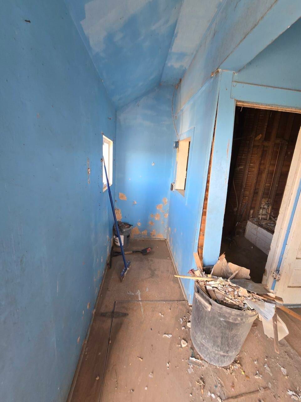 Narrow blue-painted hallway under renovation, with tools, debris, and an open doorway on the right.