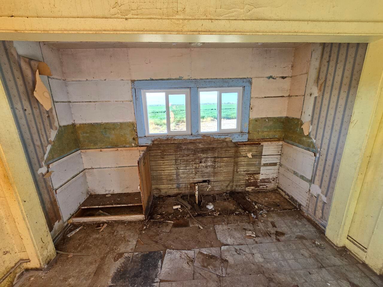 Empty, worn kitchen with peeling walls, damaged floor, and a small window above a broken sink area.