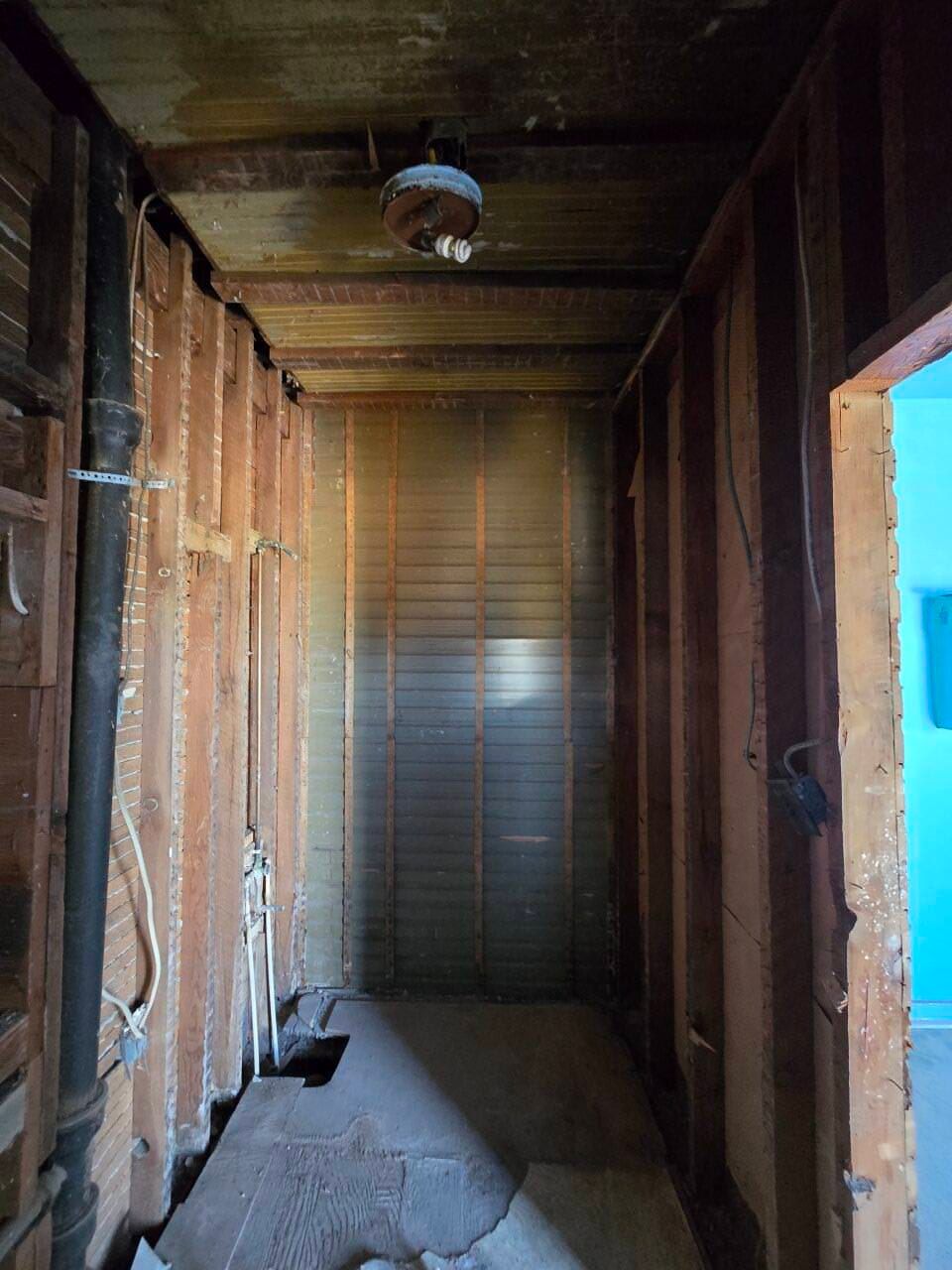 Empty unfinished hallway with exposed wooden framing and drywall, lit by daylight from a doorway.