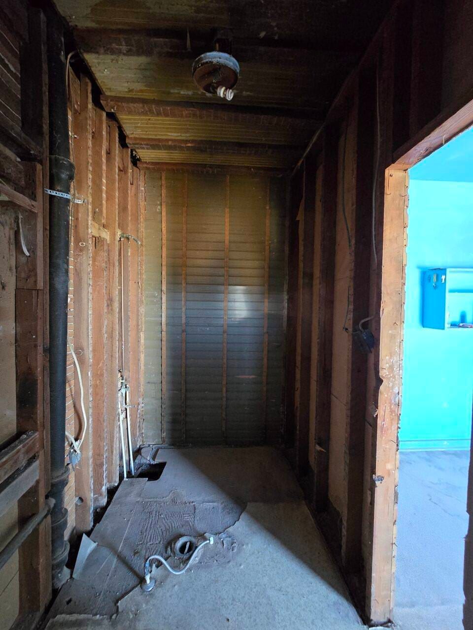 Narrow unfinished hallway with exposed wall studs, debris on the floor, and a bright blue room visible at right Narrow unfinished hallway with exposed wall studs, debris on the floor, and a bright blue room visible at right