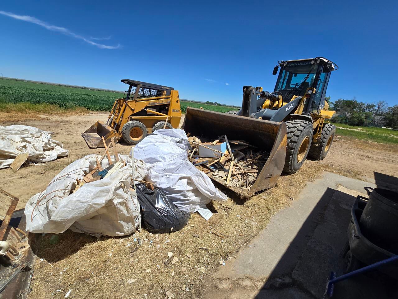 Two loaders and large bags of debris at an outdoor dirt site under a blue sky