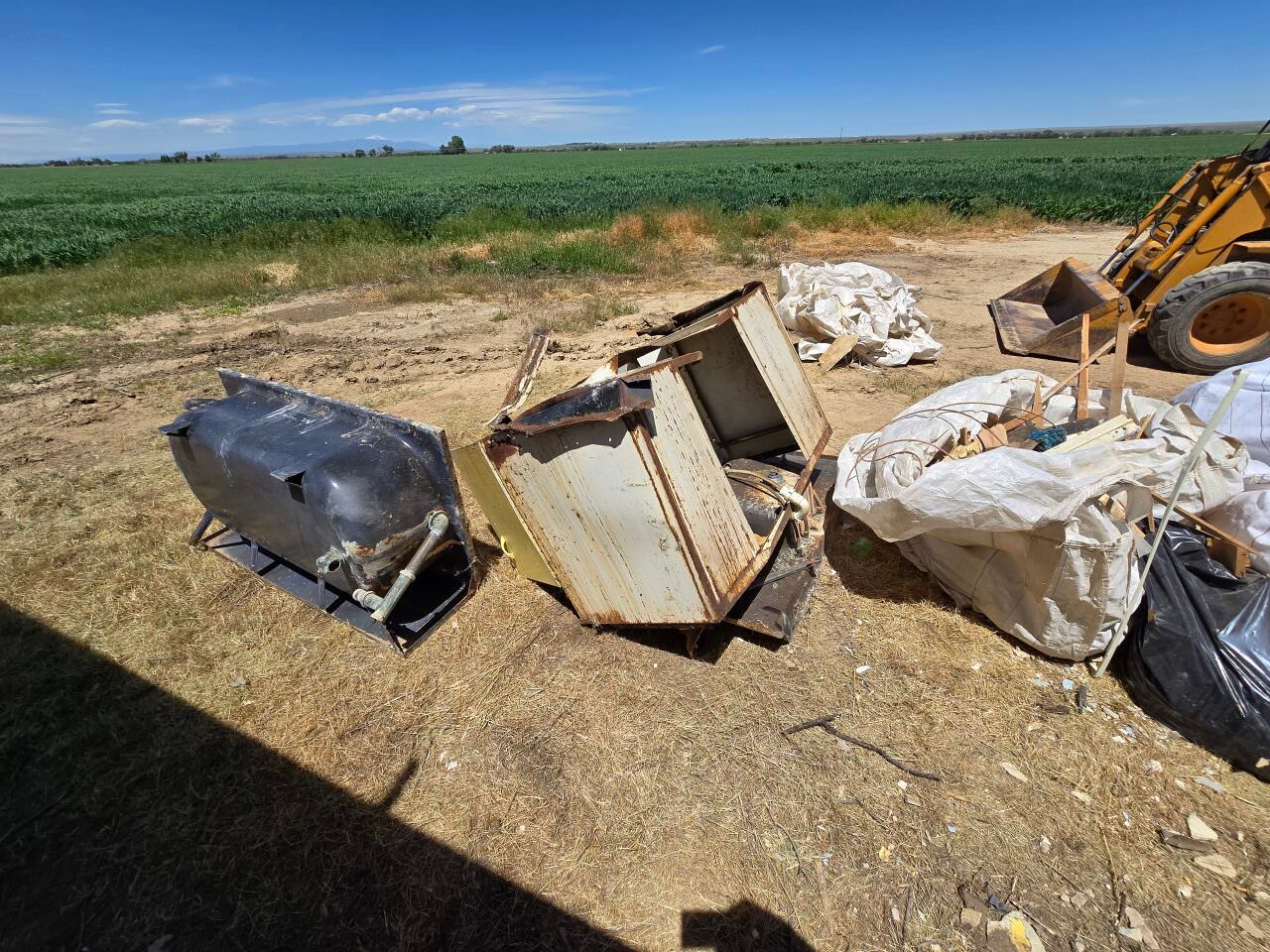 Trash and broken appliances piled on dirt beside a field under a blue sky.