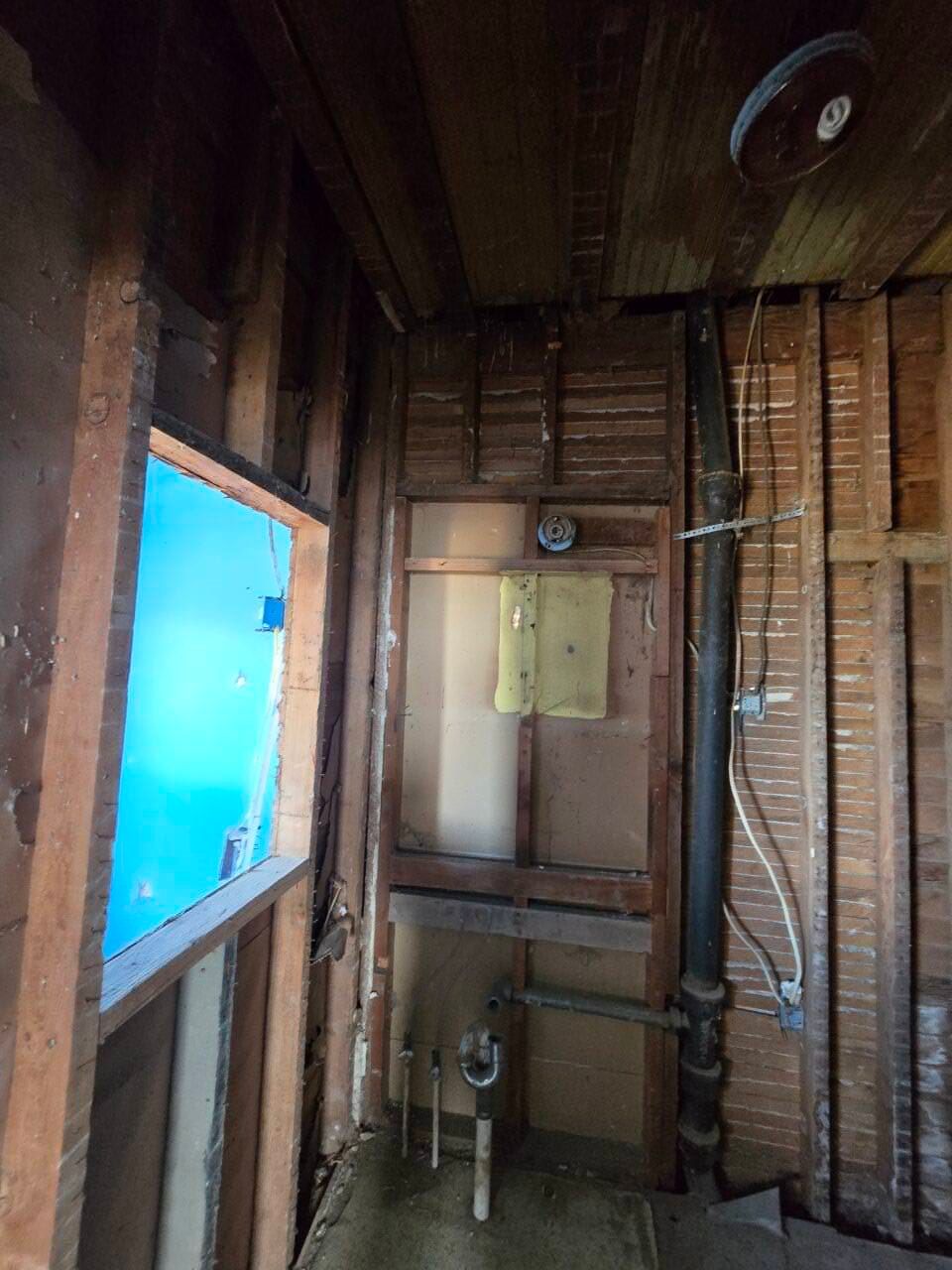 Narrow unfinished room with exposed studs, pipes, and a bright blue window opening.