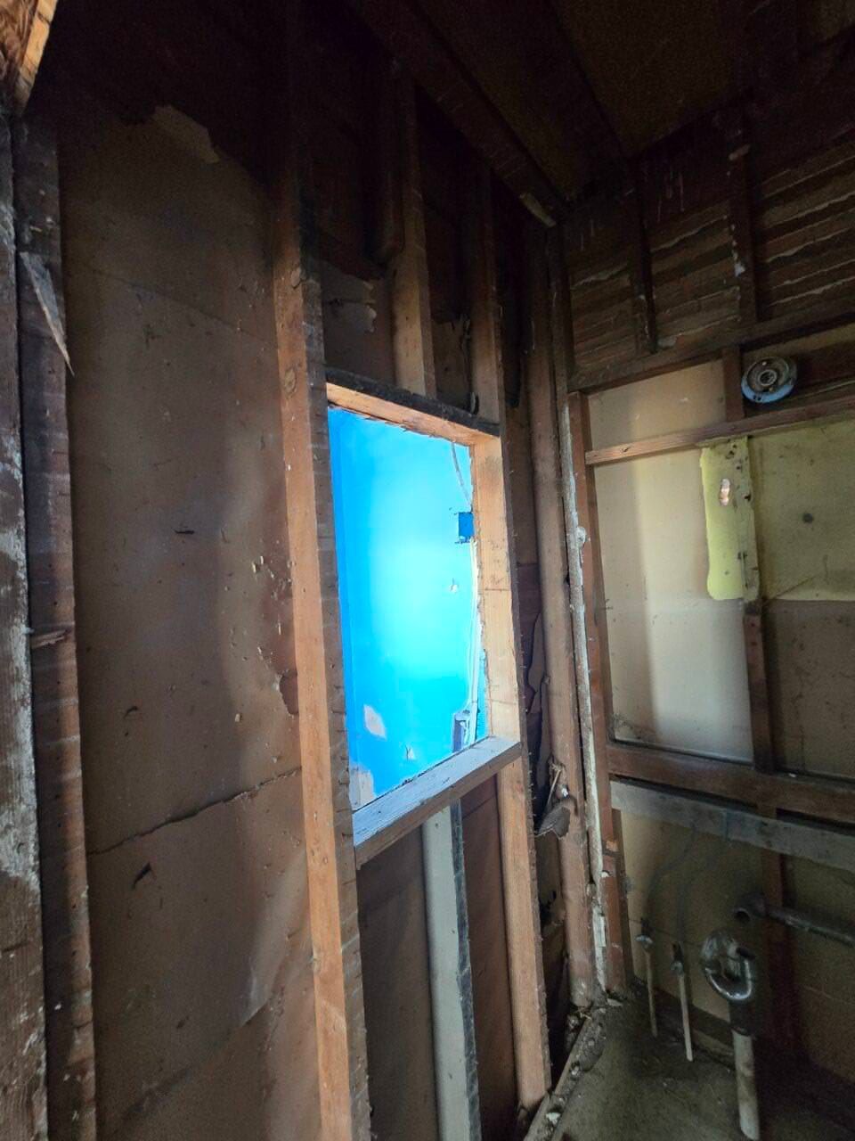 Inside a rustic wooden room, a window frames bright blue sky and rooftops outside.