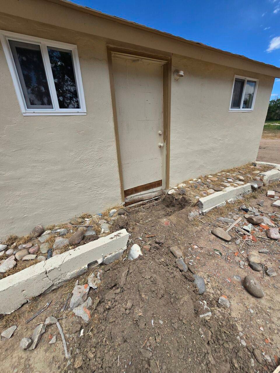Small beige building with a door and windows, surrounded by dirt and scattered rocks.