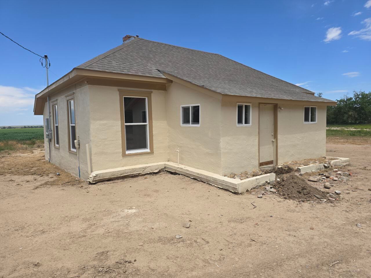 Small beige house with a gray shingled roof in a dusty, rural landscape under a blue sky