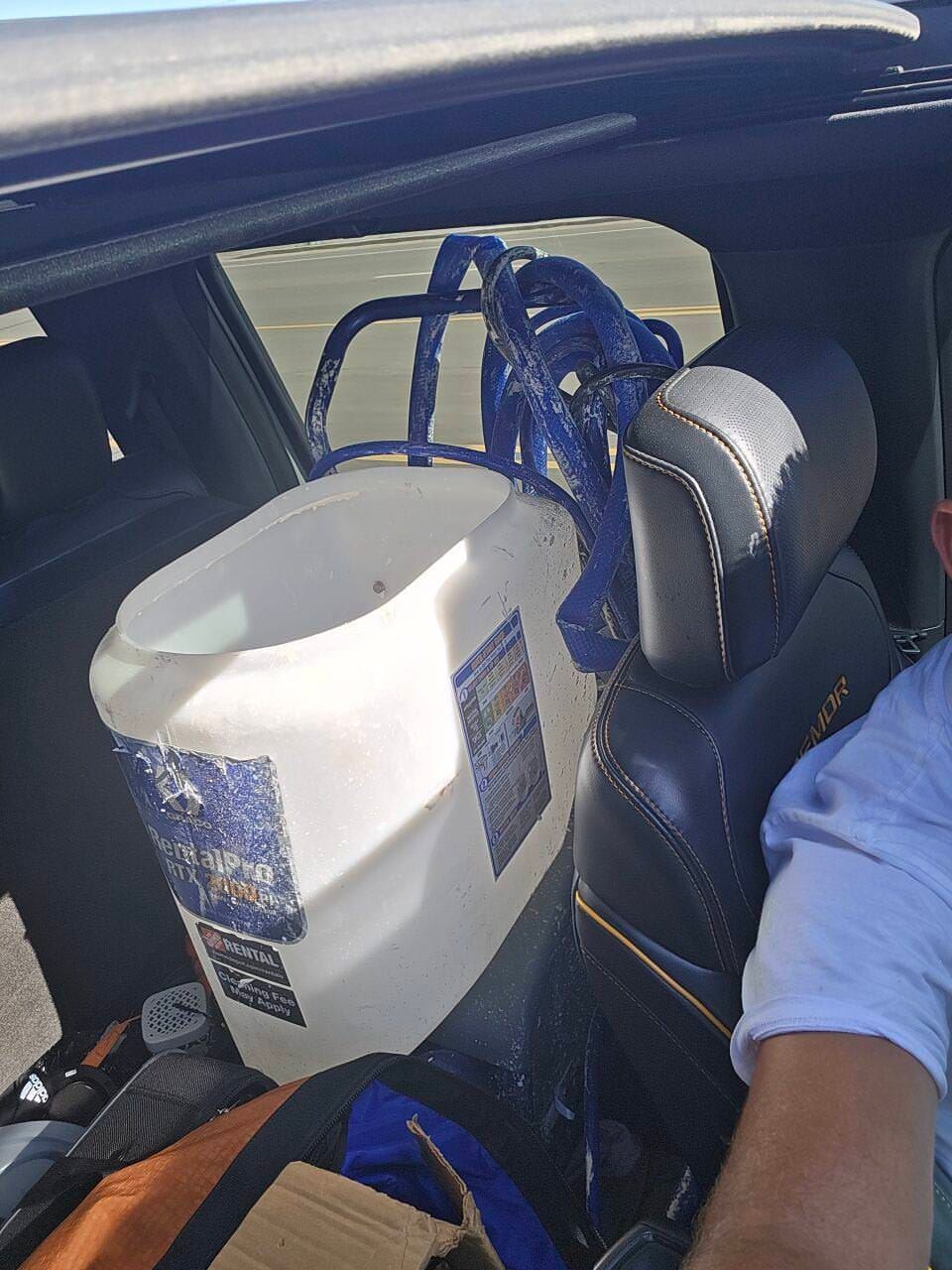 White cooler and blue backpack strapped in a car back seat beside a passenger in a blue shirt