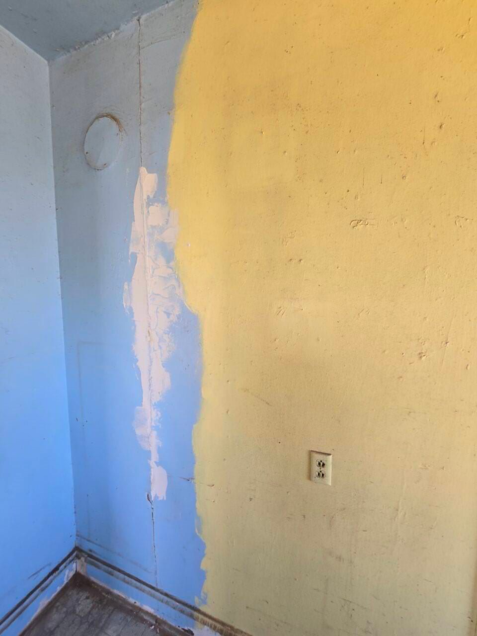 Empty room corner with blue and yellow walls, peeling paint, and a wall outlet.