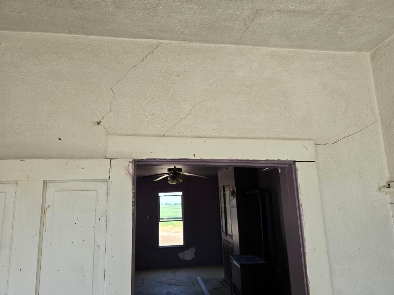 Interior doorway in a worn white room, with a view through to a darker room and a bright window beyond.