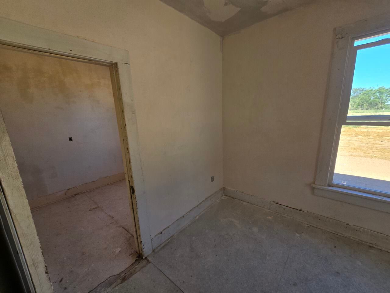 Empty unfinished room with beige walls, bare floor, open doorway, and a window overlooking the outdoors. Empty unfinished room with beige walls, bare floor, open doorway, and a window overlooking the outdoors.
