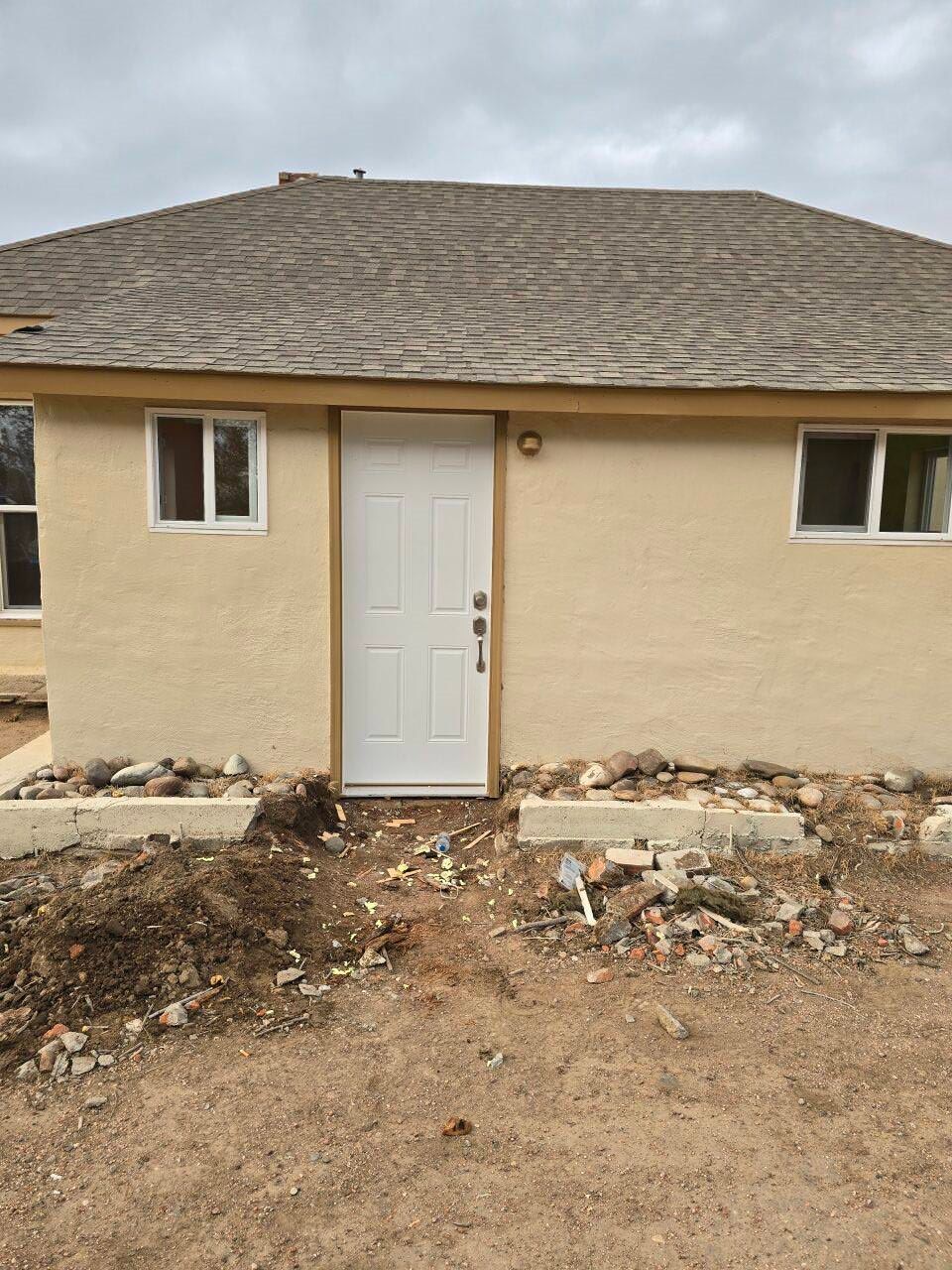 Front view of a beige house with a white door and windows, with unfinished dirt and debris in front.