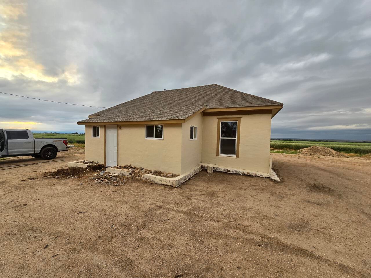 Small beige house under construction on a dirt lot beneath a cloudy sky