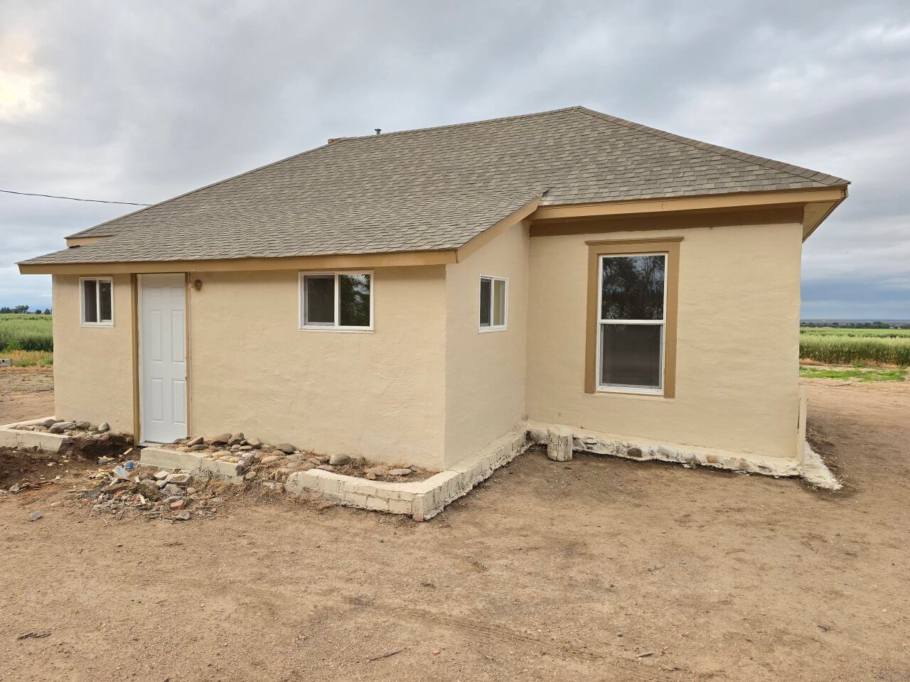 Small beige house with a shingled roof and unfinished dirt yard in a rural area